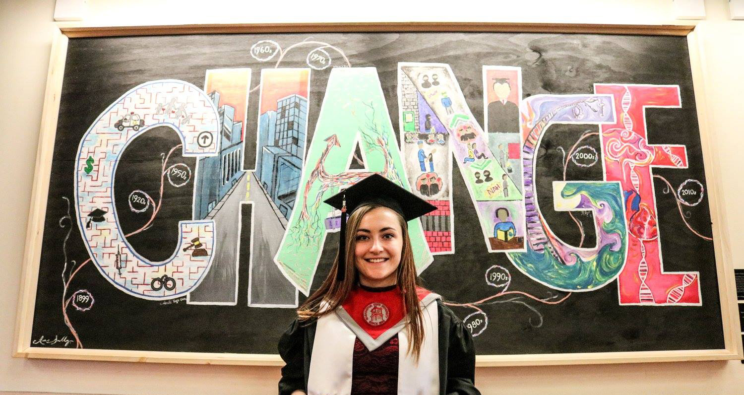 Mikala Ewert in graduation cap & gown in front of CHANGE mural.