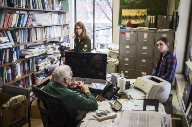 Seated professor in his office, being interviewed by two students.