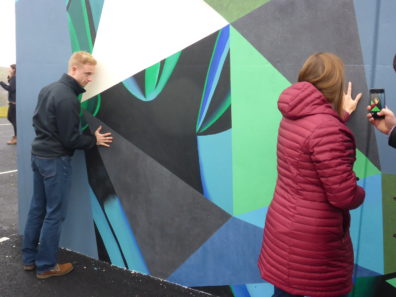 Two people put their hands against the paint on a large, outdoor mural.