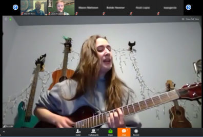 Zoom meeting screen shot of Brooke Rowland singing while playing guitar in a room with 2 guitars and a ukulele on the wall behind her.