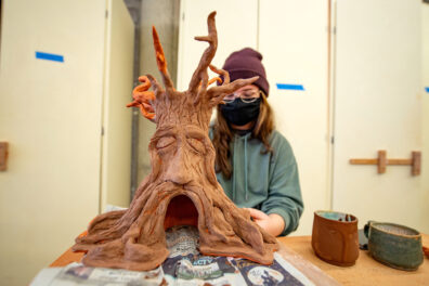 Female student working on a large clay sculpture of a tree stump with a face.