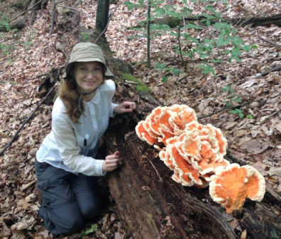 Professor Tanya Cheeke, soil microbial ecologist, kneels in a forest beside a large, orange fungus growing in an old log.