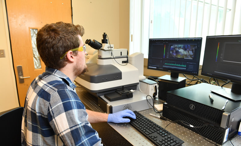 Travis Olds, in a plaid shirt, sitting at his desk in front of his computer, analyzing the chemical structure of a mineral on his computer.