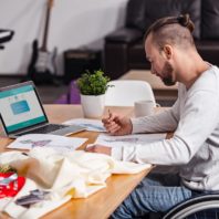 man in wheel chair studies fashion drawings at table with laptop, fabric, sewing materials