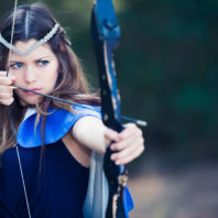 Young woman in blue costume readies to shoot arrow from bow