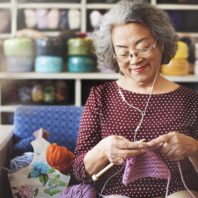 woman smiles, listens to ear buds, as she knits heavy pink yarn into garter stitch