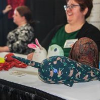 Table with stuffed narwal toy made with a blue sloth print fabric. Three women in background.