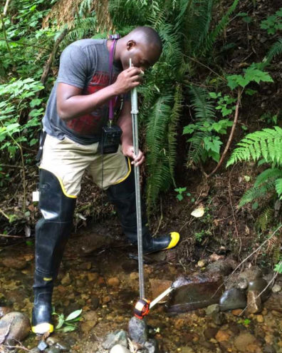 Lambert Ngenzi standing in mountain stream with water sampling tools taking water velocity measurements