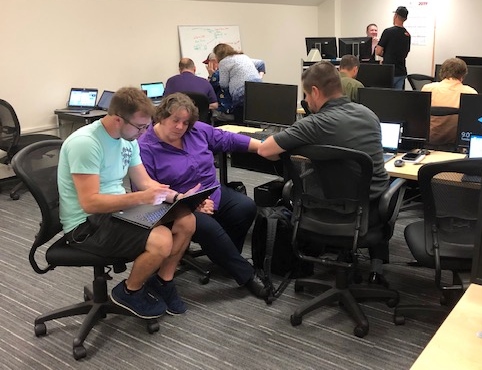 A computer lab with grey carpet, black office chairs, and beige walls. In the foreground, a woman and two men hunch over laptop computers. In the background, more people talk to each other while staring intently at computer monitors.