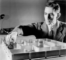 A black and white photo of a white man in a suit sitting next to a maze. He has his right hand over a robotic mice, which is in the maze