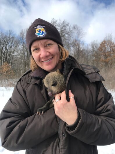 A biologist with a blond bob and brown Fish and Wildlife beanie smiles at the camera. A small bear cub is wrapped up inside her brown jacket.