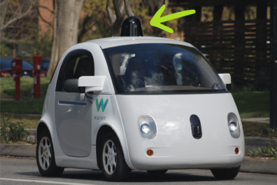 A white driverless car with the Waymo logo above its front, passenger-side tire. A green arrow points to the black, gumdrop-shaped sensor suite on top of the car.