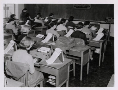 A 1960s era classroom filled with women sitting at desks and typing