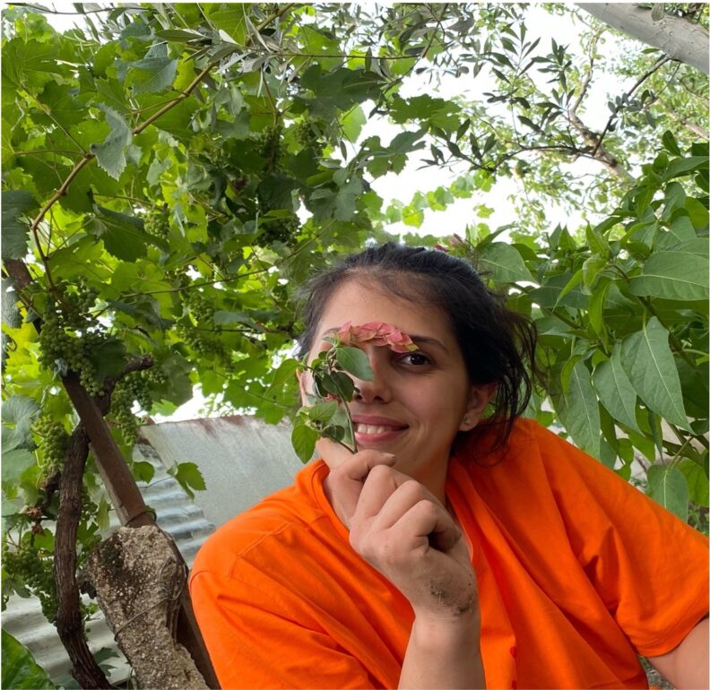 woman in orange shirt holding a flowering twig in front of her face, smiling at the camera