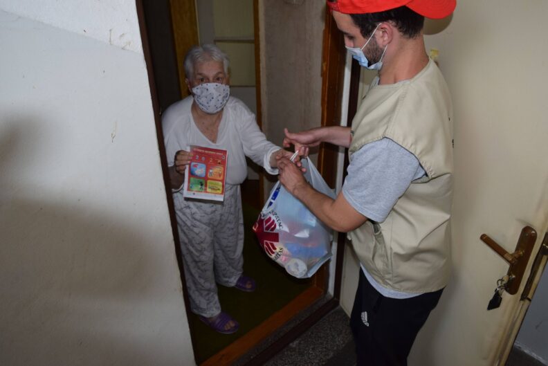 young man wearing mask handing a bag of food to an older woman at door into her house