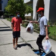 young man wearing mask offering a plastic bag holding food to a young man