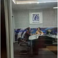 two women sitting behind glass wall talking into microphone