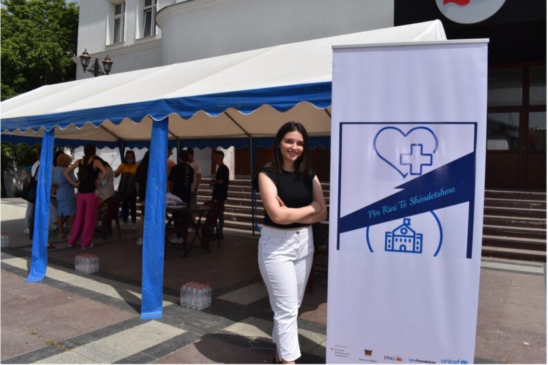 young woman standing outside at banner, sun tent behind shielding people standing on plaza