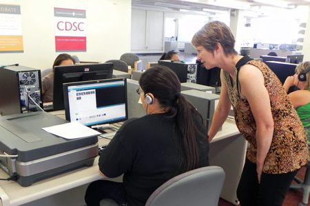 A person with their back turned working on a computer while a person is standing next to them looking over to the monitor