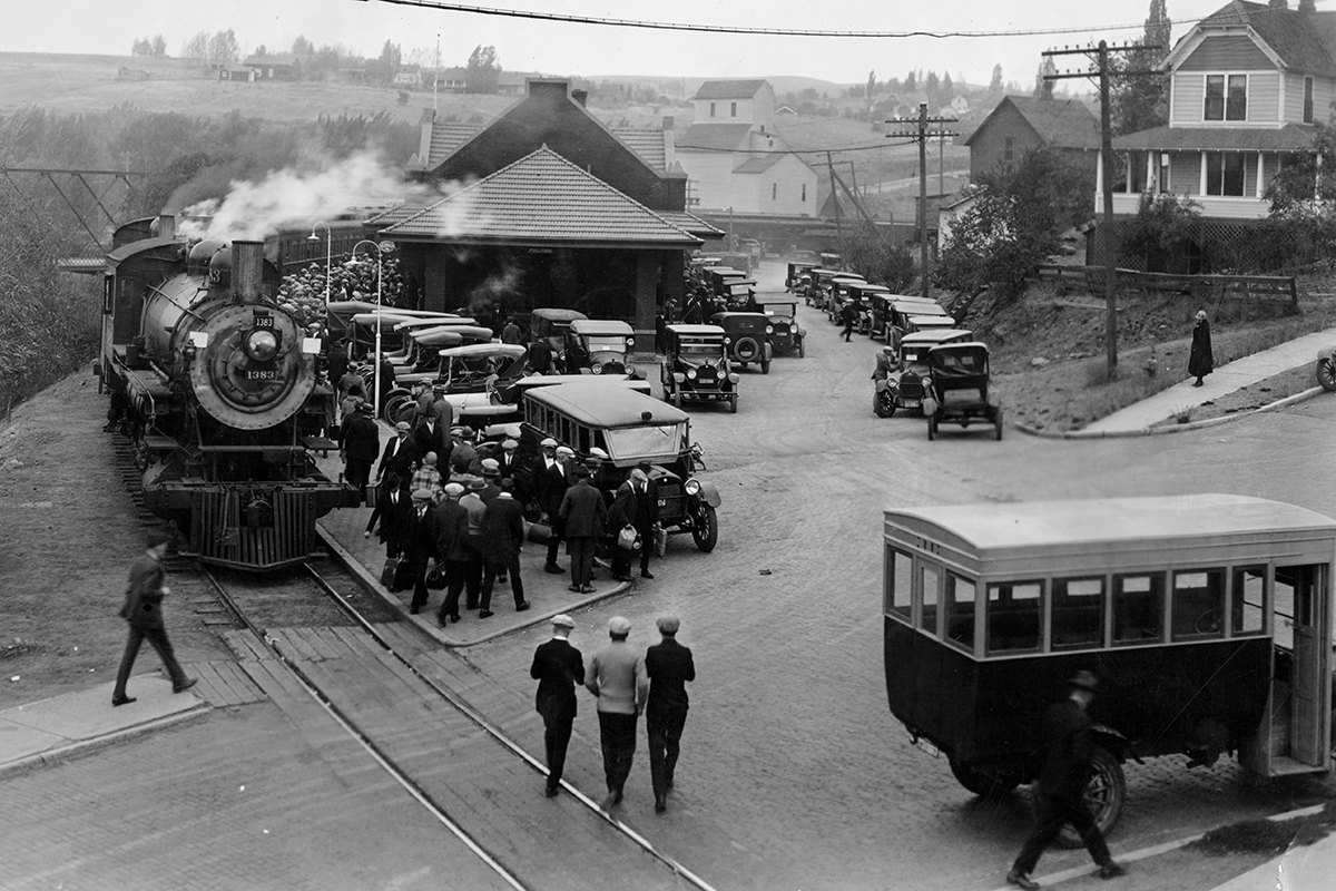 Historic photo of Pullman train depot featuring train, cars and people.