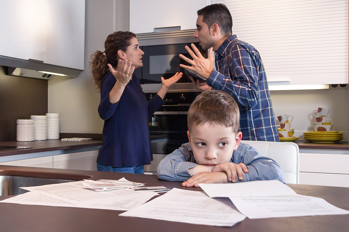 Young boy sitting at kitchen table while parents argue behind him.