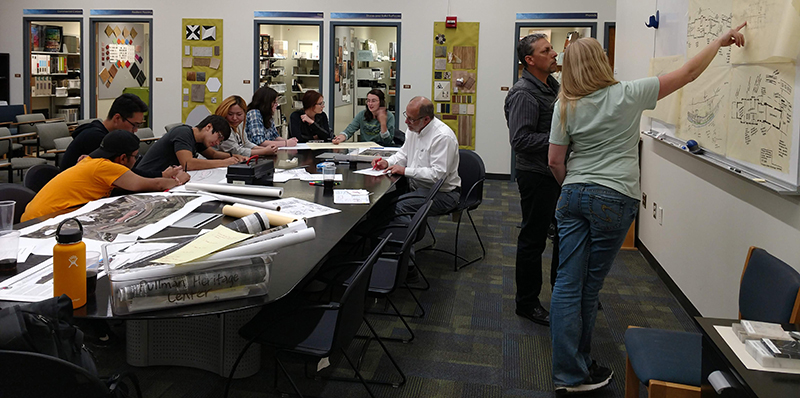 A large meeting room with students and community members seated at various tables, and lots of design papers spread out. on the right two people standing at a white board charting out ideas.