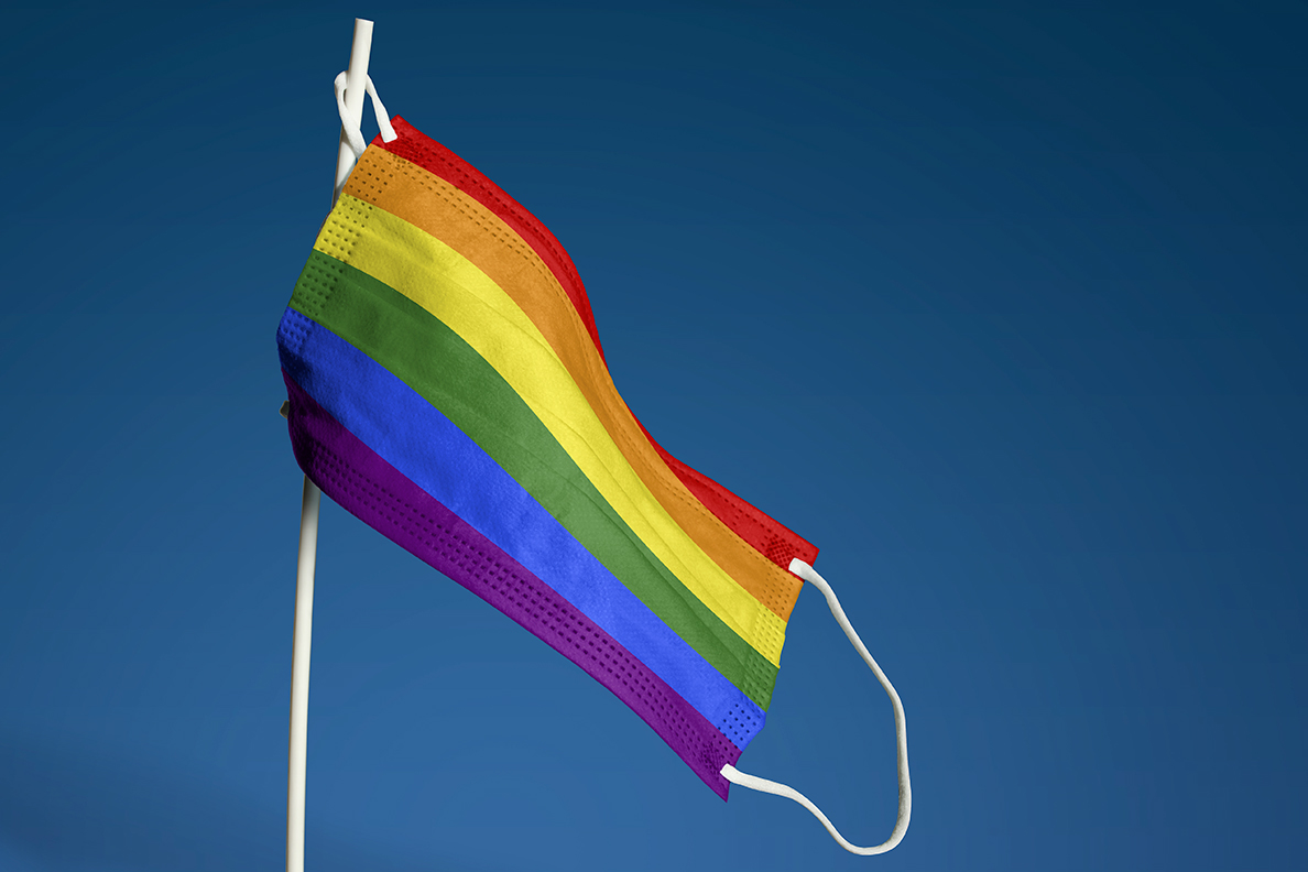 A face mask resembling an LGBTQ pride flag hanging from a stick in front of a blue background.