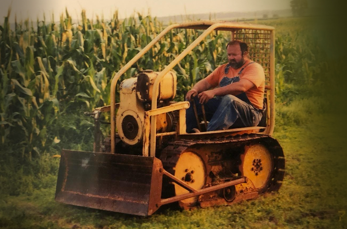 A man drives a miniature bulldozer alongside a cornfield.