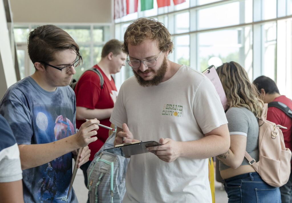 Two students with clipboards and pens talking to one another.