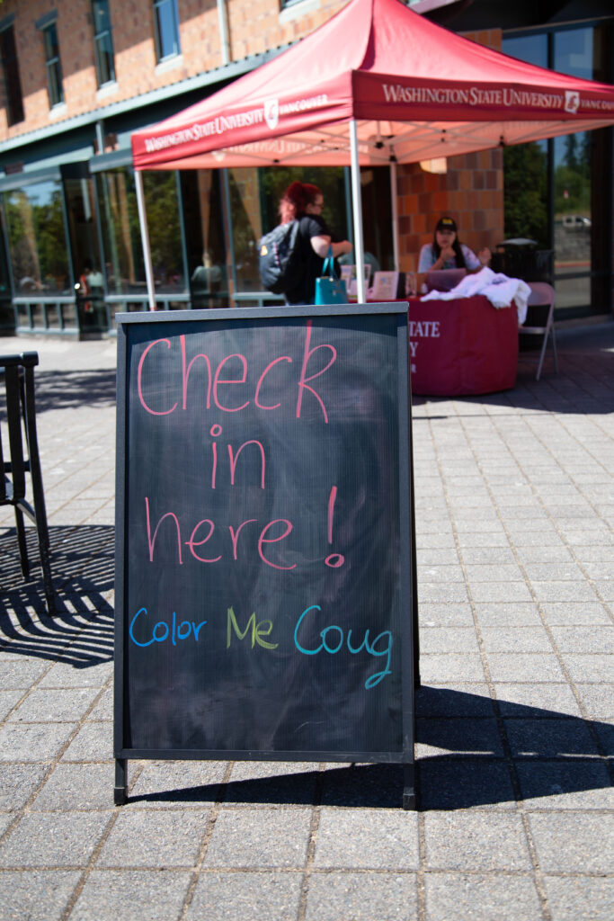A sandwich board that reads "Check in here! Color Me Coug" on the WSU Vancouver campus.