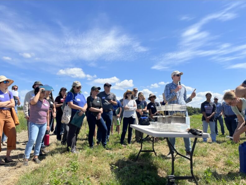 A man speaking to a group of people; demonstration equipment on a folding table in the foreground.