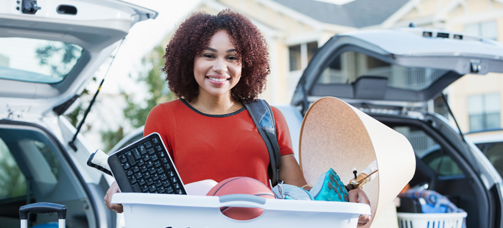 A mixed race female-presenting student smiles as she carries her personal belongings from her car to home in a laundry basket.