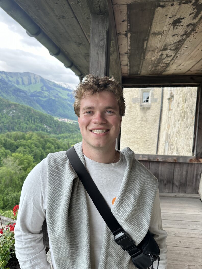 Grant Rickard while traveling, he smiles with mountains in the background.