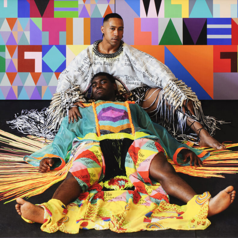 Artist Jeffrey Gibson in front of a colorful print, dressed in native art, sitting with a friend leaning on him