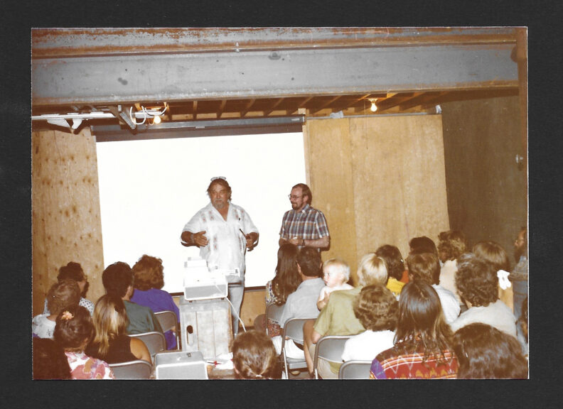 A crowd of people sit in a small room with a projector, a man presents at the front. 