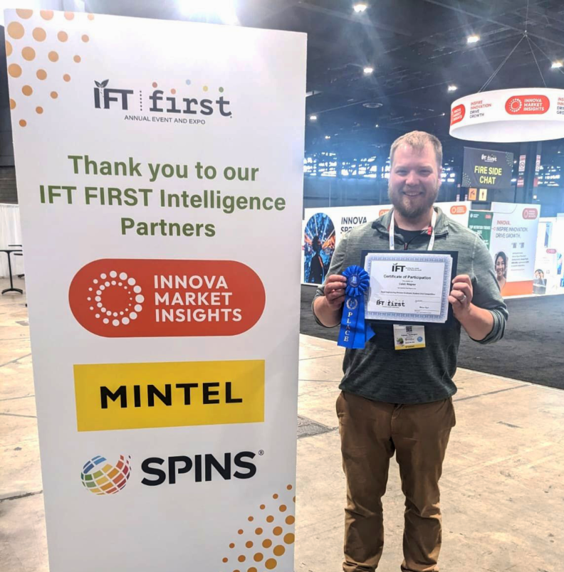 Caleb Wagner holds up a placard with a first place ribbon. He stands next to a sign that reads, "IFT First. Thank you to our IFT FIRST Intelligence Partners: Innova Market Insights, Mintel, and Spins."