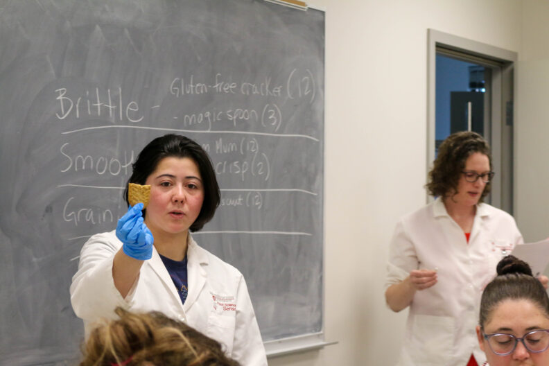 Katie holds up a cracker. Behind her is a chalkboard that says, "Brittle, Smooth, and Grain."