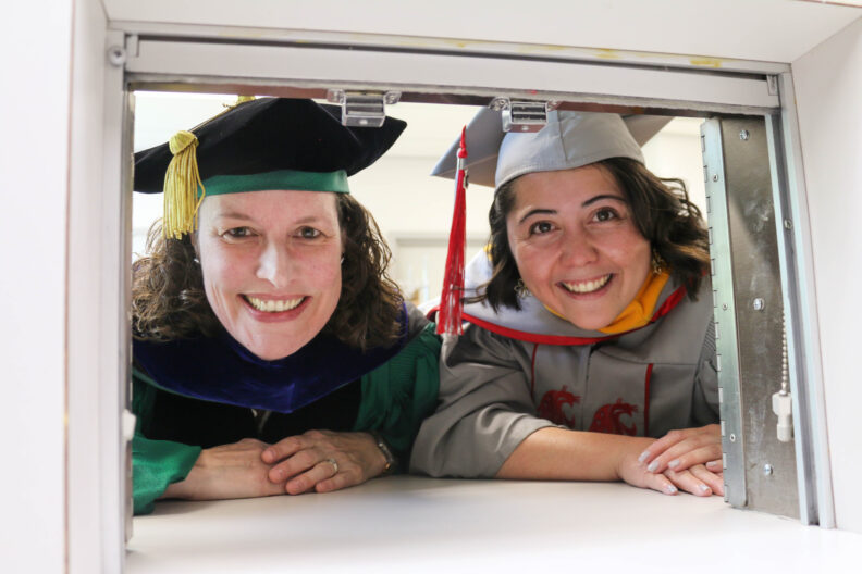Katie and Dr. Ross are in graduate and faculty regalia. They pose by one of the sensory room panel doors.