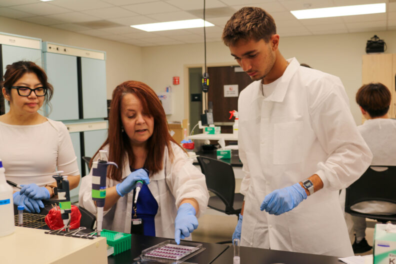 Dr. Gulhan Unlu points down at a panel with cylindrical pockets filled with a purple liquid. A student participant and Seng watch her.
