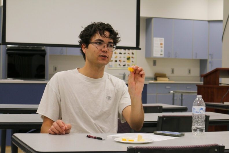 Jayden Scott holds up a square piece of cheese. There is a plate in front of him with different pieces of cheese.