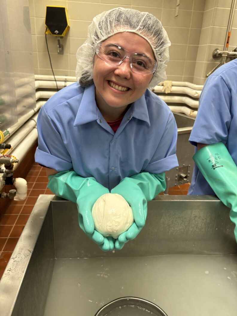 Katie wears rubber gloves, a hair net, and safety glasses. She holds a ball of mozzarella in her hands.