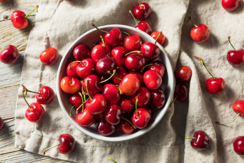 A bunch of cherries are in a bowl. Some are scattered around the bowl.