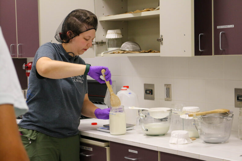 Alyssa uses a wooden spoon to stir a glass jar filled with milk and kefir grains.