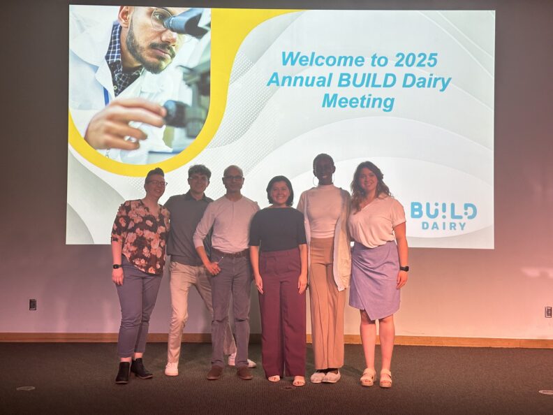 A group of students are lined up together. Behind them is a presentation slide that reads, "Welcome to 2025 Annual BUILD Dairy Meeting."