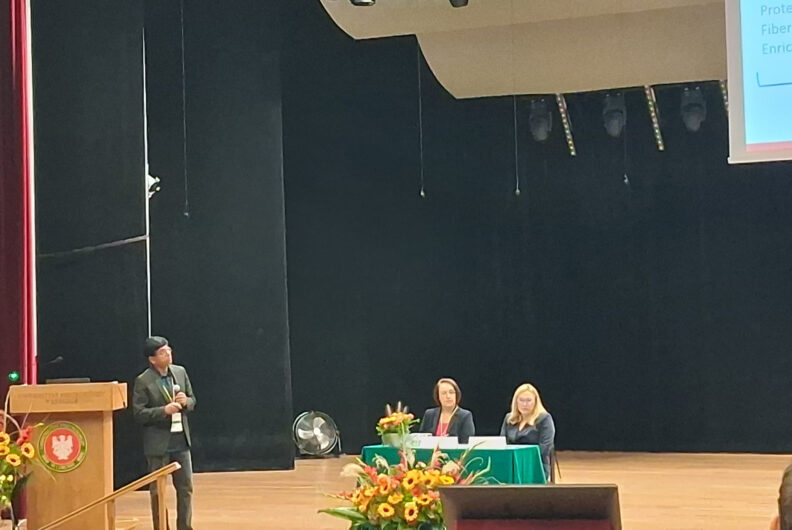Dr. Ganjyal during his presentation. Two women in formal attire are seated on a table on stage to Girish's left.