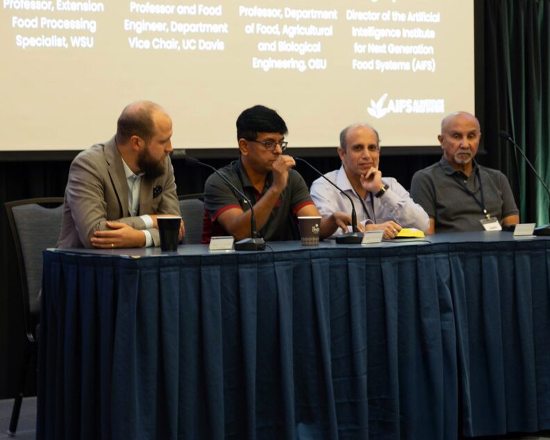 Girish Ganjyal and other panelists sit at a table on stage. Girish speaks into a microphone.