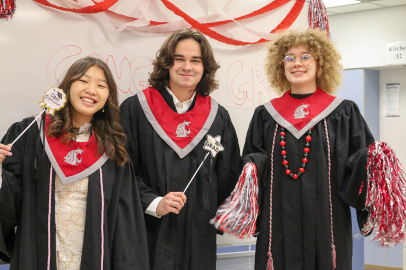 Three graduating undergrads in regalia. Two hold up wands that say "Just Graduated" and the other holds up pom poms.