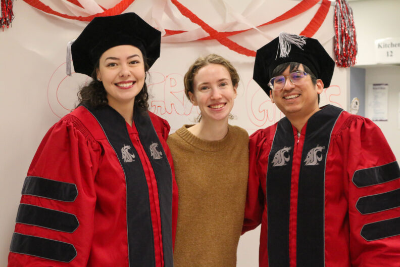Two doctorate students in graduate regalia and one student not wearing regalia.