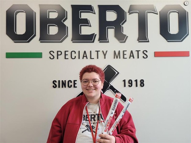 Hallie stands underneath a sign that says, "Oberto Specialist Meats Since 1918." She holds up two mega size beef sticks.