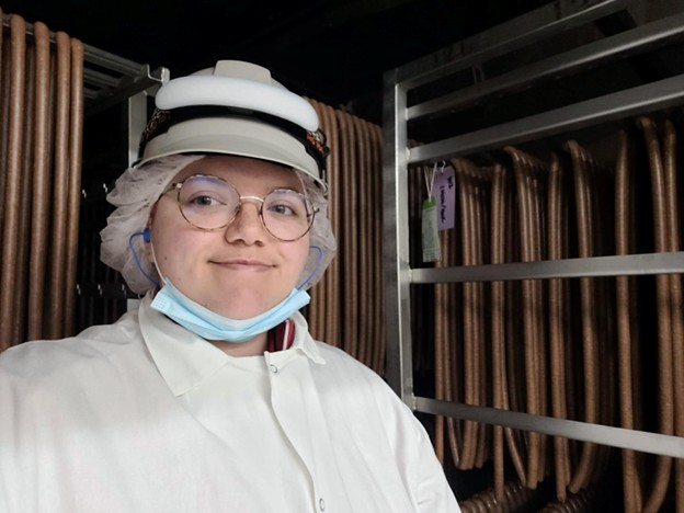 Hallie stands inside one of the smokehouses. She wears a lab coat, mask, a hair net, and a hard hat. Meat is suspended on multiple dying racks.